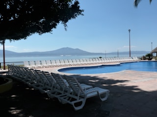 A serene pool with hydromassage jets on, surrounded by lounge chairs, adjacent to a large glass-front living room, no people present.