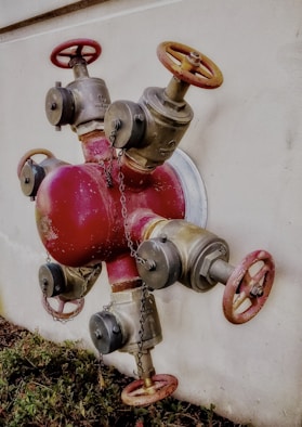 A metal valve system is mounted on a wall, featuring multiple red and silver pipes and wheel handles. The contraption includes several chains hanging around it, with grass visible at the bottom of the wall.
