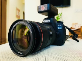 A professional Canon camera with a zoom lens attached sits on a light-colored surface. The lens cap is off, revealing the glass element with visible reflections. The camera body is prominently displaying the Canon logo and model name. In the background, there is a blurry indoor setting with some household items.