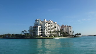 A luxurious multi-story residential building with a waterfront location, surrounded by palm trees and clear blue water. The architecture is elegant with arched windows and a light color palette.
