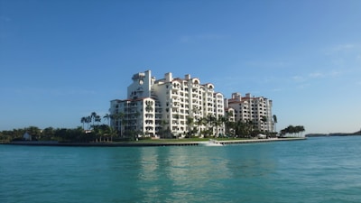 A luxurious multi-story residential building with a waterfront location, surrounded by palm trees and clear blue water. The architecture is elegant with arched windows and a light color palette.