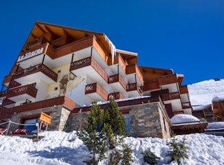 A multi-story alpine lodge with wooden balconies and stone accents, set against a clear blue sky. The building features a mural on one side and is surrounded by snow, with a few small evergreen trees in the foreground.