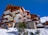 A multi-story alpine lodge with wooden balconies and stone accents, set against a clear blue sky. The building features a mural on one side and is surrounded by snow, with a few small evergreen trees in the foreground.
