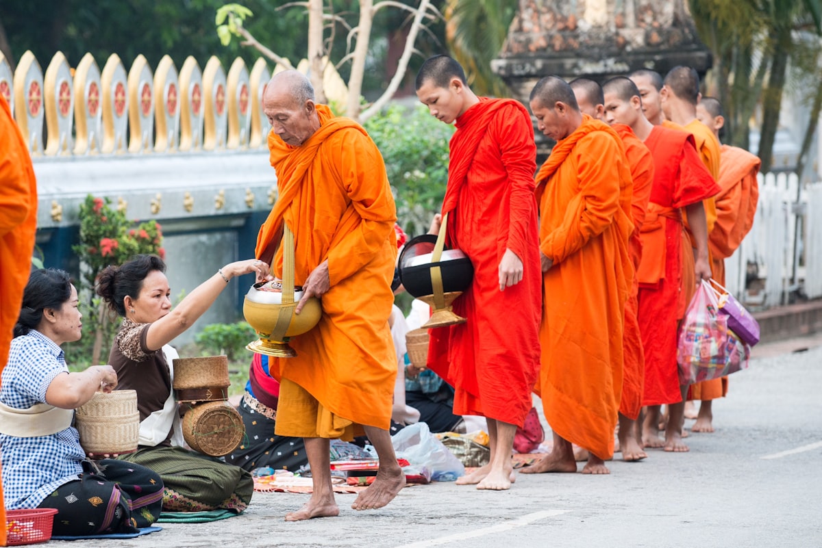Buddhist monks walking alongside villagers holding offering baskets on a quiet street in Luang Prabang