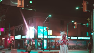 A city street at night with neon lights illuminating the area. A person in a dramatic costume stands out on the sidewalk, surrounded by various shop signs and a few people walking by.