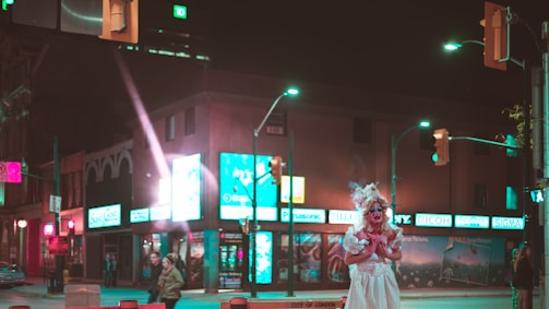 A city street at night with neon lights illuminating the area. A person in a dramatic costume stands out on the sidewalk, surrounded by various shop signs and a few people walking by.