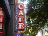 A neon sign with the word 'CAFE' in bright red and 'INTERACTIVE' in purple, mounted on a wall beside a window. The background features green leafy trees under a bright sky.
