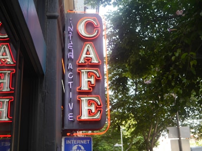 A neon sign with the word 'CAFE' in bright red and 'INTERACTIVE' in purple, mounted on a wall beside a window. The background features green leafy trees under a bright sky.