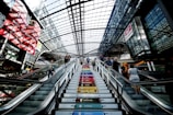 The interior of a modern, spacious building with a large, curved glass ceiling that allows natural light to flood in. An escalator and colorful stairs are visible, with people ascending and descending. The environment is busy and vibrant, with visible advertisements in the background.