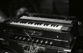 A harmonium with its keyboard and bellows visible, surrounded by knobs and wires, is placed on a dark surface. The lighting suggests a dimly lit environment, focusing on the instrument.