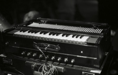 A harmonium with its keyboard and bellows visible, surrounded by knobs and wires, is placed on a dark surface. The lighting suggests a dimly lit environment, focusing on the instrument.