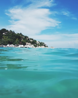 Calm ocean water with gentle waves in the foreground and a coastline lined with houses and lush green vegetation in the distance under a bright blue sky with scattered white clouds.