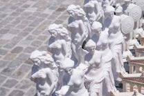 A collection of white statues resembling classical figures, possibly representing Greek or Roman women in a market or outdoor setting. The statues are aligned in rows, showcasing intricate details in their hair and garments. The ground surface appears to be made of stone tiles.