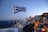A Greek flag is prominently displayed in the foreground, blowing in the wind. In the background, there is a scenic view of a coastal town with iconic white-washed buildings and blue domes. The town is illuminated with warm lights as dusk settles in, and the sun is setting over the horizon. The sea is visible in the distance, with a boat sailing on it.