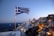 A Greek flag is prominently displayed in the foreground, blowing in the wind. In the background, there is a scenic view of a coastal town with iconic white-washed buildings and blue domes. The town is illuminated with warm lights as dusk settles in, and the sun is setting over the horizon. The sea is visible in the distance, with a boat sailing on it.