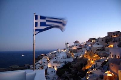 A Greek flag is prominently displayed in the foreground, blowing in the wind. In the background, there is a scenic view of a coastal town with iconic white-washed buildings and blue domes. The town is illuminated with warm lights as dusk settles in, and the sun is setting over the horizon. The sea is visible in the distance, with a boat sailing on it.