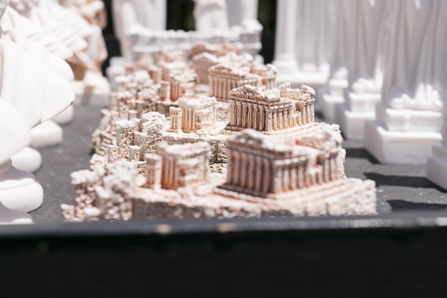 A series of small, intricate architectural models resembling ancient ruins or temples, crafted with detailed columns and stone-like textures. The models are displayed on a dark surface, surrounded by other statues or sculptures.