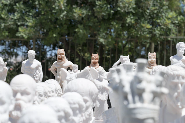 Numerous white statues are displayed outdoors with a background of green foliage and a fence. The statues depict a range of figures, including warriors, mythical creatures, and people in various poses. Some statues have features reminiscent of classical Greek or Roman sculptures, while others appear to be more fantastical with additional color details.