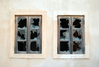 Two old wooden-framed windows with multiple broken panes set in a faded wall. The glass is shattered in a pattern suggesting damage and neglect.