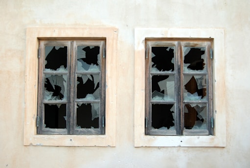 Two old wooden-framed windows with multiple broken panes set in a faded wall. The glass is shattered in a pattern suggesting damage and neglect.