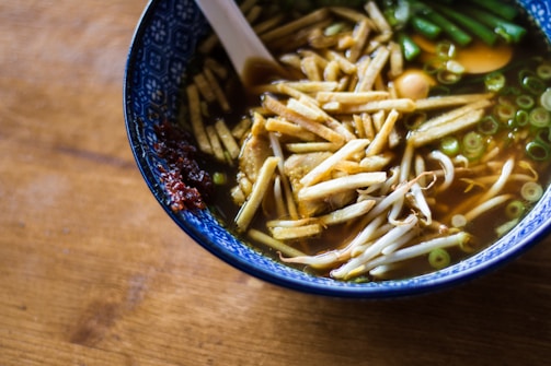 A ceramic bowl filled with soup, containing bean sprouts, sliced green onions, bamboo shoots, and a side of chili paste. The soup has a light brown broth and is served with a white ceramic spoon. The bowl rests on a wooden surface and features an intricate blue and white pattern on the rim.