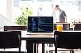 A laptop displaying code rests on a wooden table in a bright cafe. Beside the laptop, a glass of orange juice with a straw adds a pop of color. Sunlight streams in through large windows, creating a warm and inviting atmosphere. In the background, a blurred figure appears to be walking, with green plants adding a touch of nature to the setting.