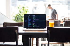 A laptop displaying code rests on a wooden table in a bright cafe. Beside the laptop, a glass of orange juice with a straw adds a pop of color. Sunlight streams in through large windows, creating a warm and inviting atmosphere. In the background, a blurred figure appears to be walking, with green plants adding a touch of nature to the setting.
