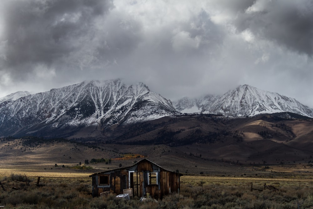 Cabina di legno marrone abbandonata al centro della valle con vista sulla catena montuosa innevata sotto la formazione di nuvole grigie durante il giorno