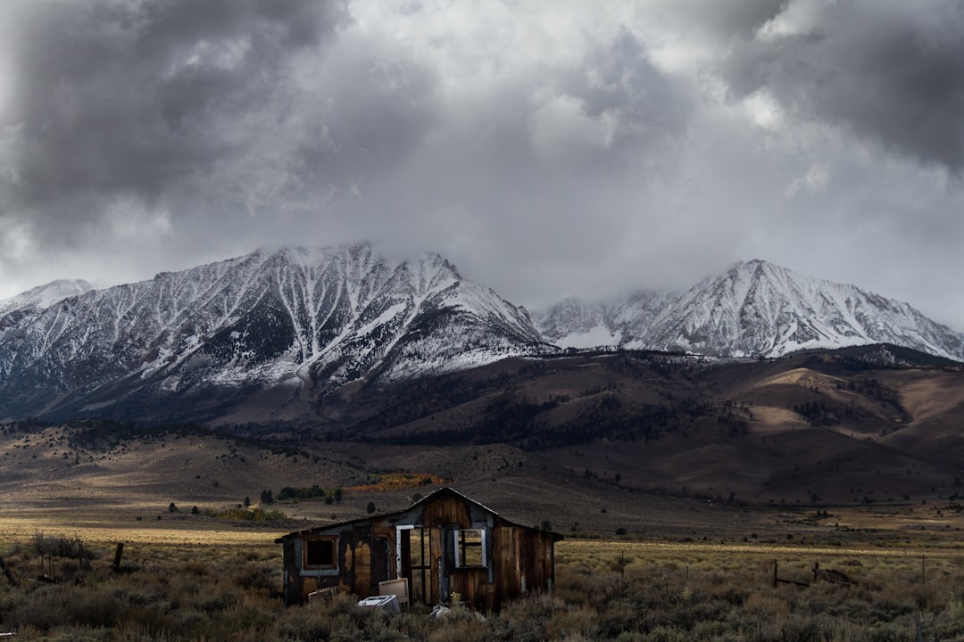 abandoned brown wooden cabin at middle of valley with view of snow-covered mountain range under gray cloud formation during daytime