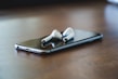 A modern smartphone and wireless earbuds displayed on a wooden desk with soft ambient lighting.