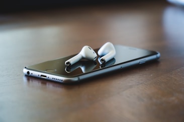 A smartphone is placed on a wooden surface with a pair of wireless earbuds resting on top. The earbuds contrast the sleek, dark screen of the phone.
