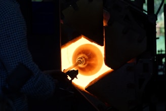 A person is engaged in the process of glassblowing, manipulating a glowing piece of glass inside a furnace. The furnace emits a bright orange light, casting shadows on the surrounding equipment and the person.