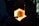 A person is engaged in the process of glassblowing, manipulating a glowing piece of glass inside a furnace. The furnace emits a bright orange light, casting shadows on the surrounding equipment and the person.