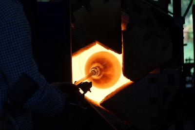 A person is engaged in the process of glassblowing, manipulating a glowing piece of glass inside a furnace. The furnace emits a bright orange light, casting shadows on the surrounding equipment and the person.
