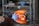 A glassblowing process is taking place, with a glowing molten glass piece being shaped by a torch. Flames and heat are visible, indicating high temperatures. The background shows shelves with tools and materials, suggesting a workshop environment.