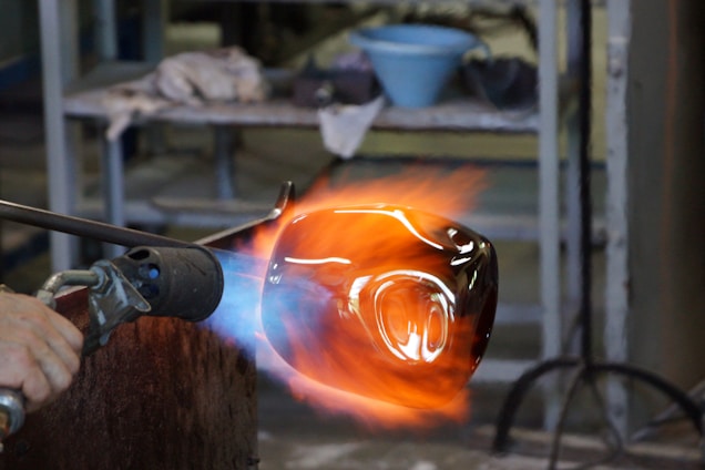 A glassblowing process is taking place, with a glowing molten glass piece being shaped by a torch. Flames and heat are visible, indicating high temperatures. The background shows shelves with tools and materials, suggesting a workshop environment.