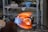 A glassblowing process is taking place, with a glowing molten glass piece being shaped by a torch. Flames and heat are visible, indicating high temperatures. The background shows shelves with tools and materials, suggesting a workshop environment.
