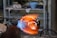 A glassblowing process is taking place, with a glowing molten glass piece being shaped by a torch. Flames and heat are visible, indicating high temperatures. The background shows shelves with tools and materials, suggesting a workshop environment.