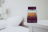 A glass bottle of Dolce & Gabbana perfume resting on a white bedside table. The bottle features a gradient design with a dark base transitioning to a lighter amber tone at the top. In the background, a blurred view of a white bed and a lamp adds to the minimalistic and elegant setting.