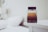 A glass bottle of Dolce & Gabbana perfume resting on a white bedside table. The bottle features a gradient design with a dark base transitioning to a lighter amber tone at the top. In the background, a blurred view of a white bed and a lamp adds to the minimalistic and elegant setting.