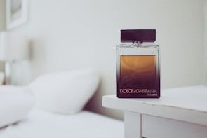 A glass bottle of Dolce & Gabbana perfume resting on a white bedside table. The bottle features a gradient design with a dark base transitioning to a lighter amber tone at the top. In the background, a blurred view of a white bed and a lamp adds to the minimalistic and elegant setting.