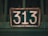 A vintage, weathered metal sign displays the number 313. The sign is dark green in color with large white numerals, and it shows signs of rust and wear. The background is a blurred shade of green, suggesting it is attached to an old gate or structure.