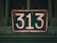 A vintage, weathered metal sign displays the number 313. The sign is dark green in color with large white numerals, and it shows signs of rust and wear. The background is a blurred shade of green, suggesting it is attached to an old gate or structure.