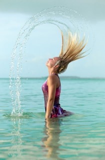 A person with long blonde hair is in a colorful bathing suit, knee-deep in clear turquoise water. They are flipping their hair back, creating an arc of water droplets in the air.