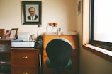 A home office setup with a black office chair in front of a wooden desk and under a large framed portrait of a man. The desk has a few stationery items and a small lamp. A printer is placed on a wooden cabinet next to the desk. The room is softly lit by daylight coming through a window on the right.