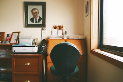 A home office setup with a black office chair in front of a wooden desk and under a large framed portrait of a man. The desk has a few stationery items and a small lamp. A printer is placed on a wooden cabinet next to the desk. The room is softly lit by daylight coming through a window on the right.