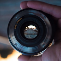 A hand holding a camera lens is directing the view towards a railway track that is visible through the lens' center. The focus is on the track, with the surrounding areas blurred, creating a tunnel effect through the lens.