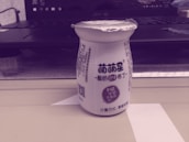 A small bottle with printed text on it stands on a shelf or table. The bottle has a foil top and Chinese characters on its label, indicating it might be a dairy or yogurt drink. Behind the bottle, there is an open laptop partially visible.