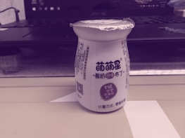 A small bottle with printed text on it stands on a shelf or table. The bottle has a foil top and Chinese characters on its label, indicating it might be a dairy or yogurt drink. Behind the bottle, there is an open laptop partially visible.