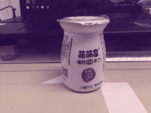 A small bottle with printed text on it stands on a shelf or table. The bottle has a foil top and Chinese characters on its label, indicating it might be a dairy or yogurt drink. Behind the bottle, there is an open laptop partially visible.